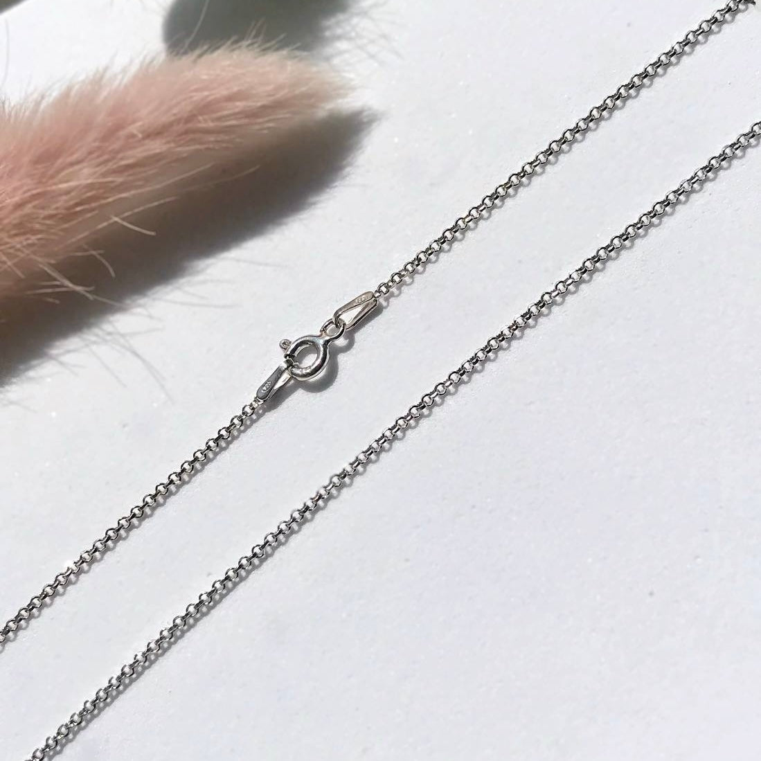 A silver necklace with a length of 50 cm, displayed on a white surface with a pink feather to the left and a quality assurance stamp to the right.